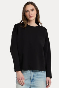 A woman wearing a black long-sleeve crewneck sweater and light wash blue jeans, standing against a plain light gray studio background.