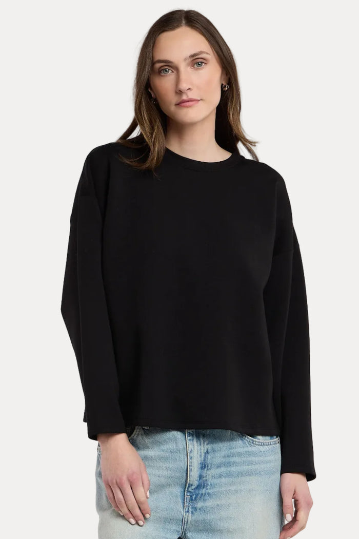 A woman wearing a black long-sleeve crewneck sweater and light wash blue jeans, standing against a plain light gray studio background.