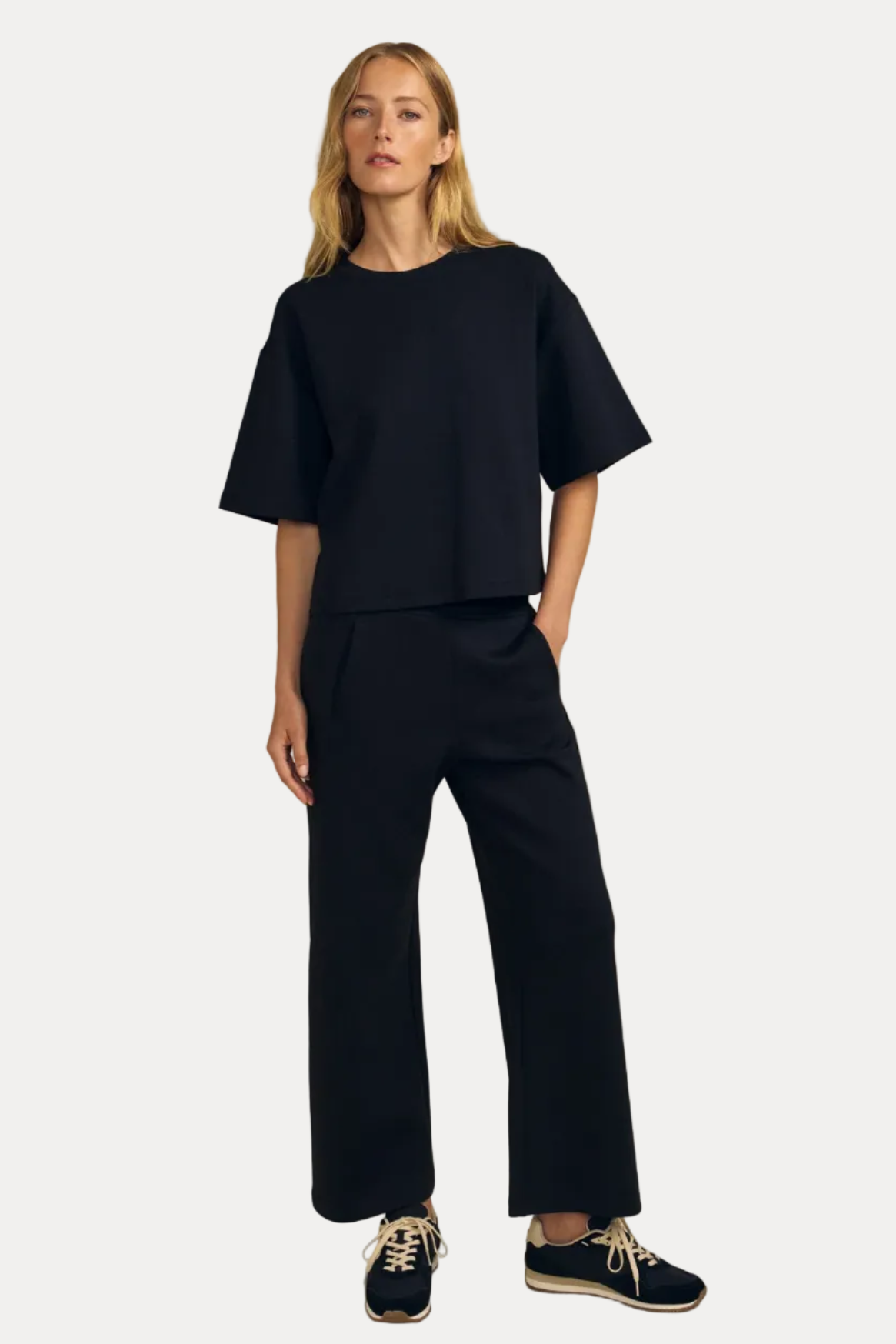 A woman modeling a black short-sleeve cropped top and matching black wide-leg trousers, paired with navy sneakers, against a solid beige studio background.
