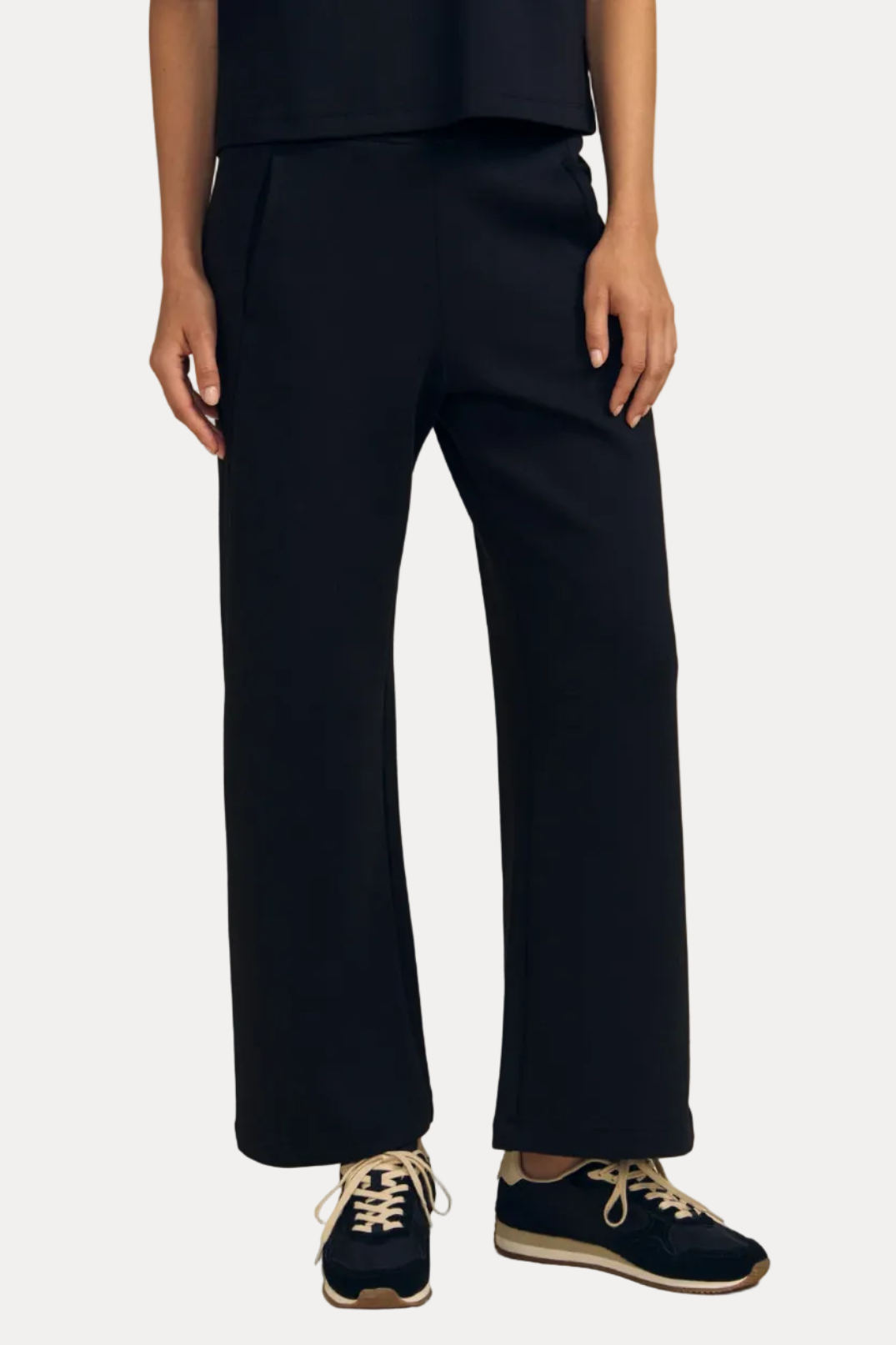 A model from the waist down wearing dark navy wide-leg trousers, a matching dark navy top, and navy sneakers with beige laces, set against a solid beige studio background.