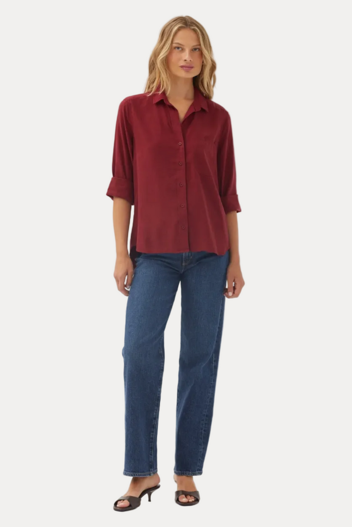 A woman modeling a burgundy button-down shirt with three-quarter sleeves and blue jeans against a plain light gray studio background.