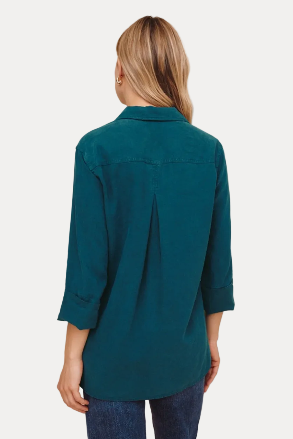 A woman viewed from the back wearing a teal long-sleeve button-up shirt and blue jeans against a plain beige studio background.