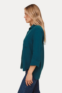 A woman shown in side profile wearing a teal button-up shirt with three-quarter sleeves and dark blue jeans, set against a solid light beige background.
