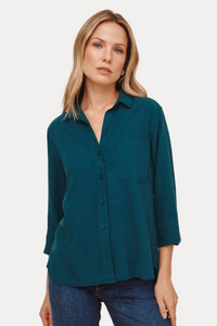 A woman wearing a teal long-sleeve button-up shirt and blue jeans, standing against a solid beige studio background.