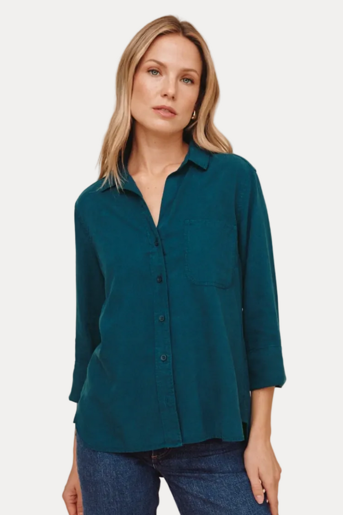 A woman wearing a teal long-sleeve button-up shirt and blue jeans, standing against a solid beige studio background.