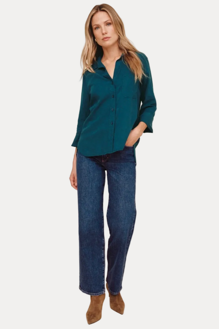 A woman modeling a teal button-down shirt with dark blue jeans and brown suede boots, standing against a solid beige studio background.