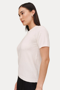 A woman shown in side profile wearing a fitted short-sleeve white knit top and dark pants against a plain warm beige studio background.