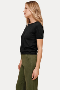A woman in profile wearing a short-sleeve black knit top and olive green pants, standing against a plain beige studio background.