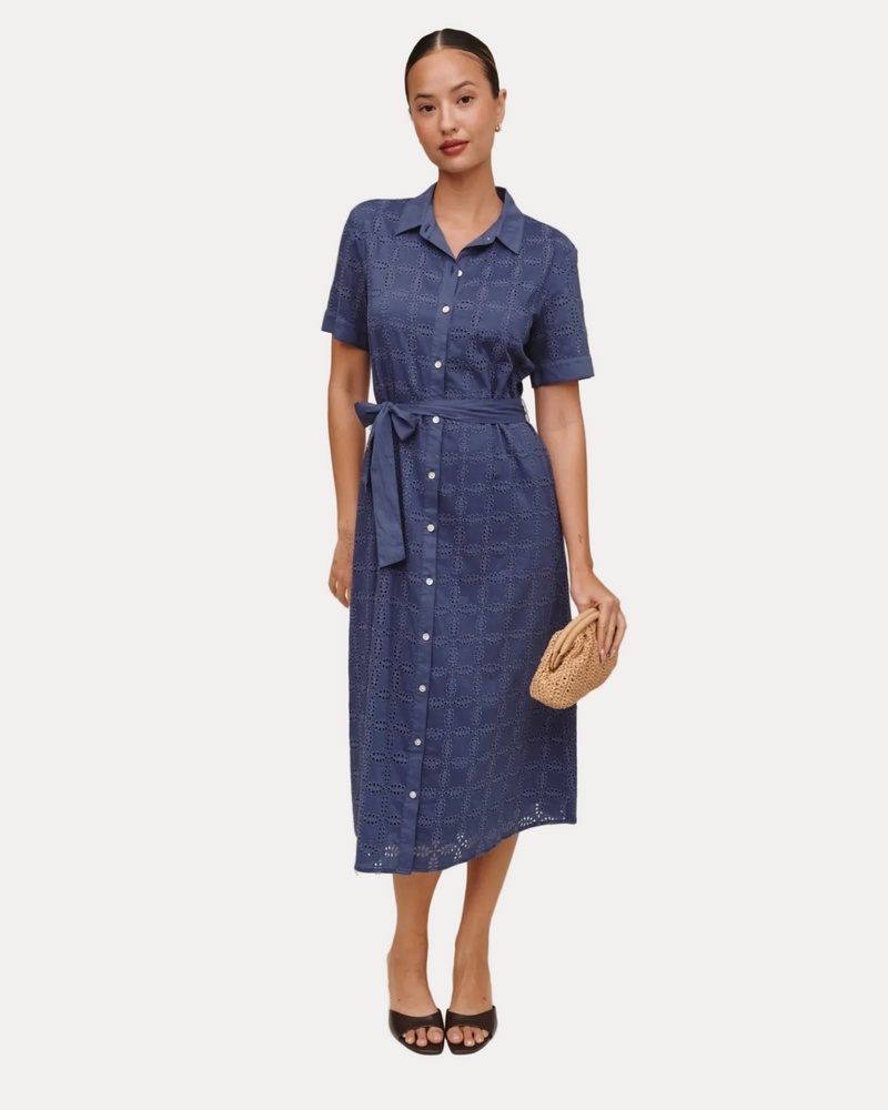 A woman modeling a short-sleeve blue midi shirt dress with a button-down front, collar, and matching tie belt, standing against a plain beige studio background.