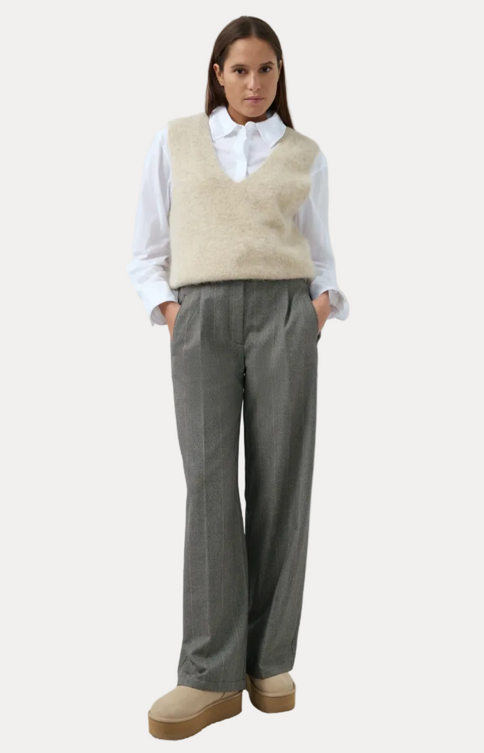 A woman stands against a light blue studio background wearing a white collared shirt, a cream fuzzy sleeveless vest, gray wide-leg trousers, and beige platform slip-on shoes.