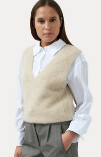 A woman wearing a fuzzy beige knit vest over a white collared shirt and gray trousers, photographed against a light blue studio background.