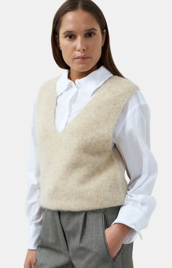 A woman wearing a fuzzy beige knit vest over a white collared shirt and gray trousers, photographed against a light blue studio background.