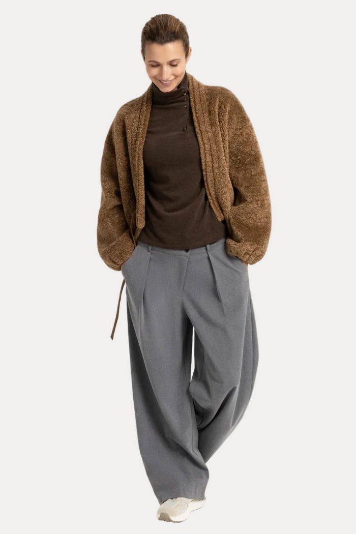 A woman modeling a brown textured knit cardigan over a dark brown turtleneck, paired with wide-leg gray trousers and white sneakers, against a plain white studio background.