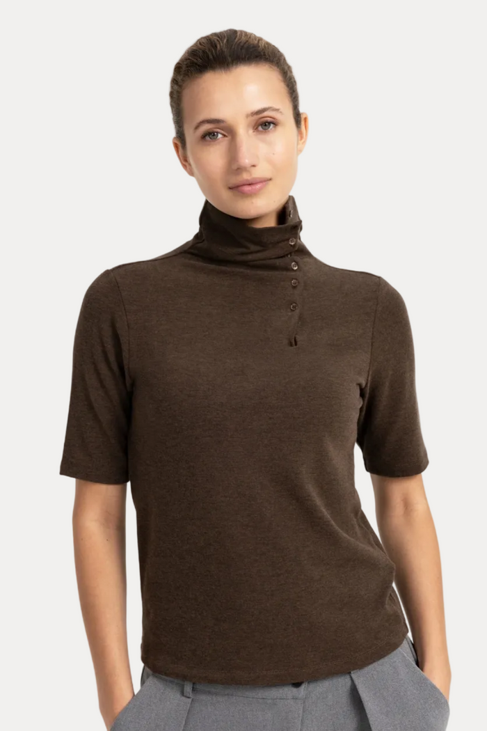 A woman modeling a short-sleeve brown turtleneck top with a buttoned placket, paired with gray trousers, against a plain white studio background.