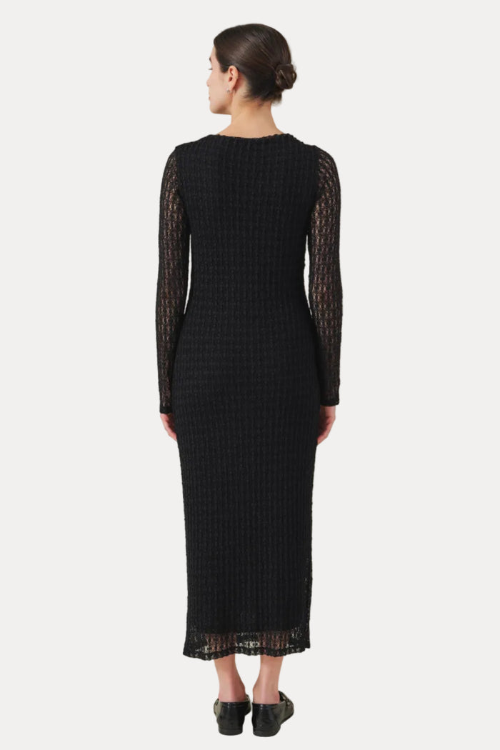 A woman viewed from the back wearing a black long-sleeve midi dress with textured knit fabric and sheer lace sleeves, paired with black shoes, against a plain light beige studio background.