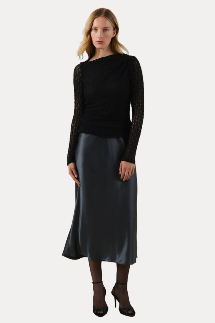 A woman stands against a plain warm beige studio background wearing a black textured knit long-sleeve top with sheer patterned sleeves and a dark satin midi skirt.