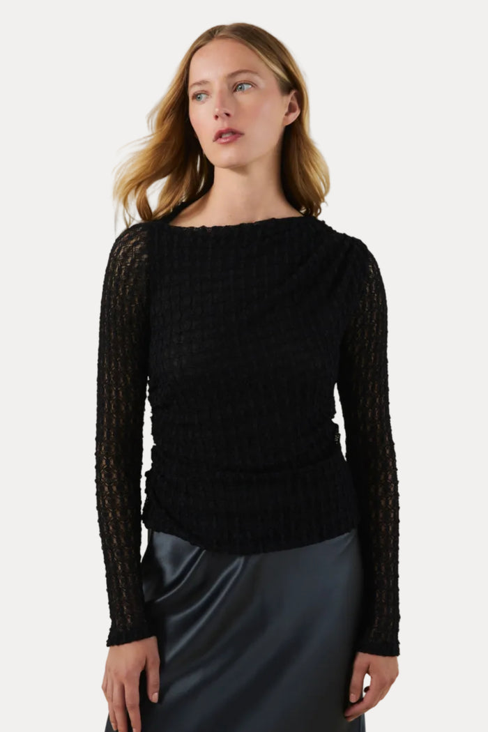 A woman modeling a black textured knit long-sleeve top with a boat neckline, paired with a dark satin skirt, against a plain beige studio background.