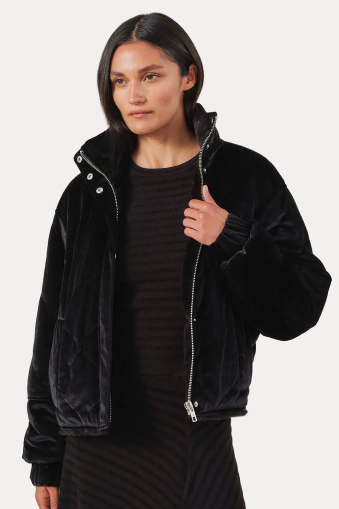 A model wearing a black velvet zip-up jacket over a dark ribbed top and skirt, photographed against a solid light beige background.