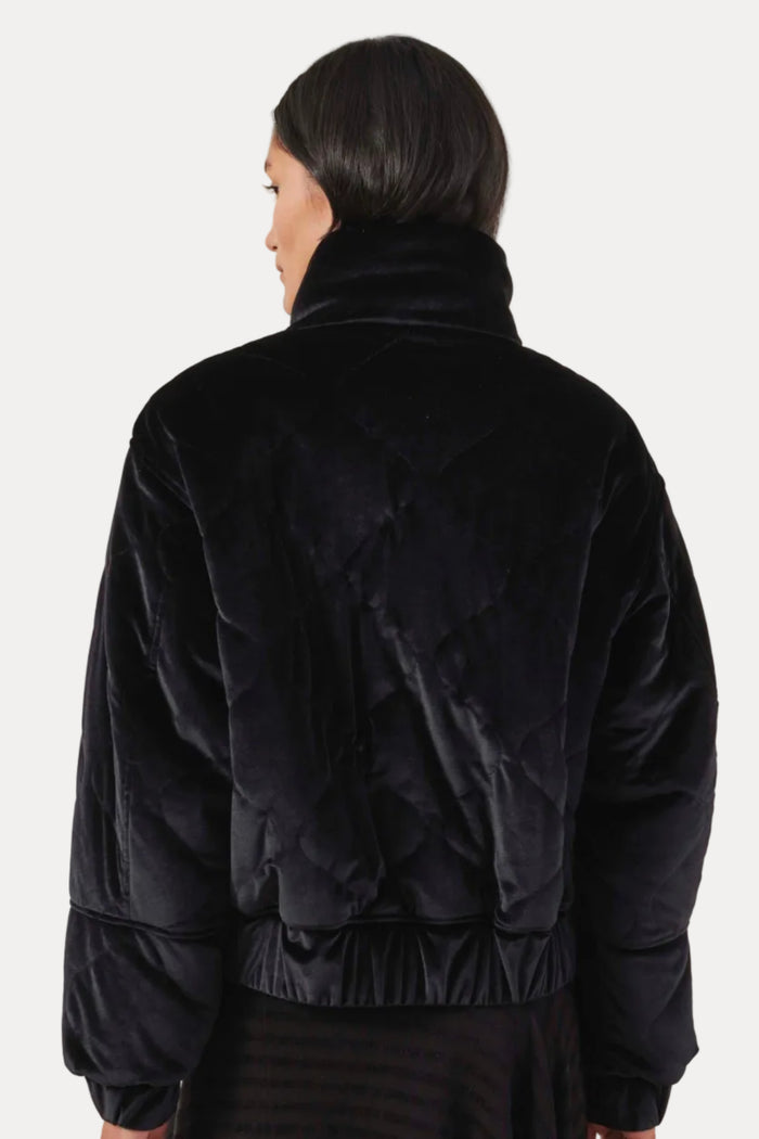 A person shown from the back wearing a black quilted velvet jacket with a high collar, photographed against a plain light beige studio background.