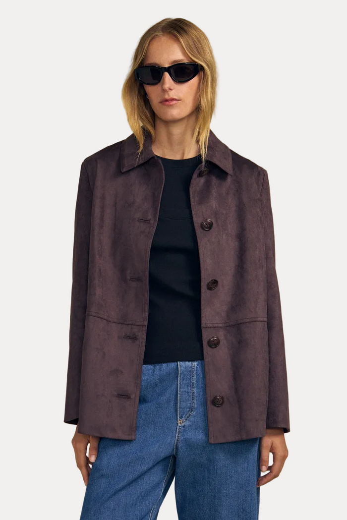 A model wearing a dark brown suede button-up blazer over a black top and blue jeans, set against a solid beige background.