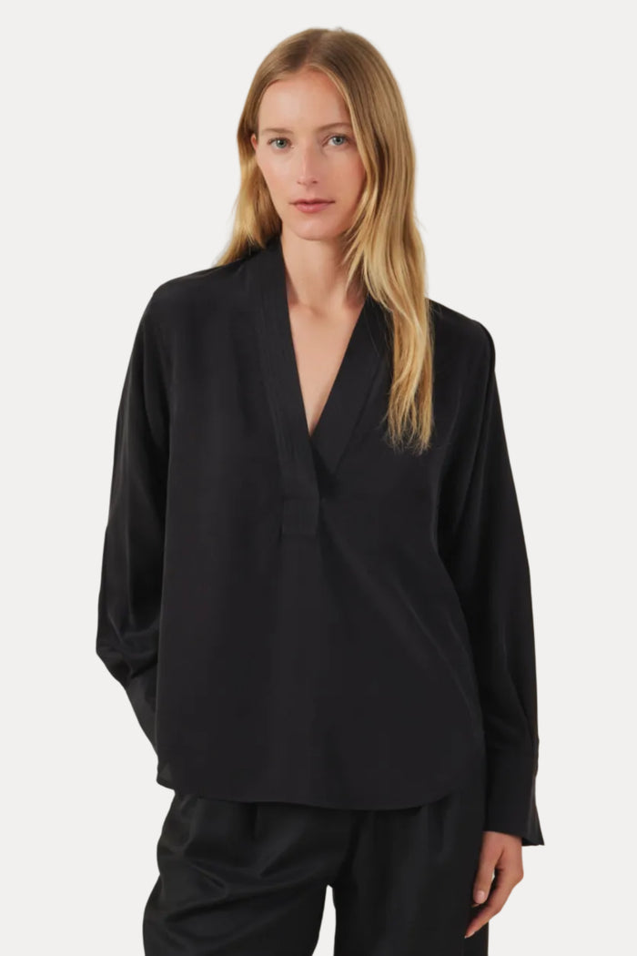 A woman modeling a black long-sleeve wrap-style blouse with a deep V-neck and black wide-leg trousers, photographed against a plain light beige studio background.