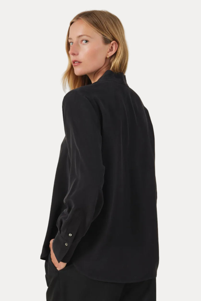 A woman wearing a black long-sleeve button-up shirt and matching black trousers, shown from a side-back angle against a plain light beige studio background.
