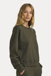 A woman modeling a dark olive green crewneck sweatshirt and matching sweatpants against a solid beige background.