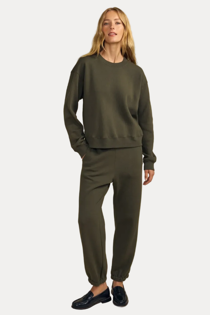 A woman modeling a matching olive green lounge set consisting of a crewneck sweatshirt and jogger pants, paired with dark loafers, against a solid beige studio background.